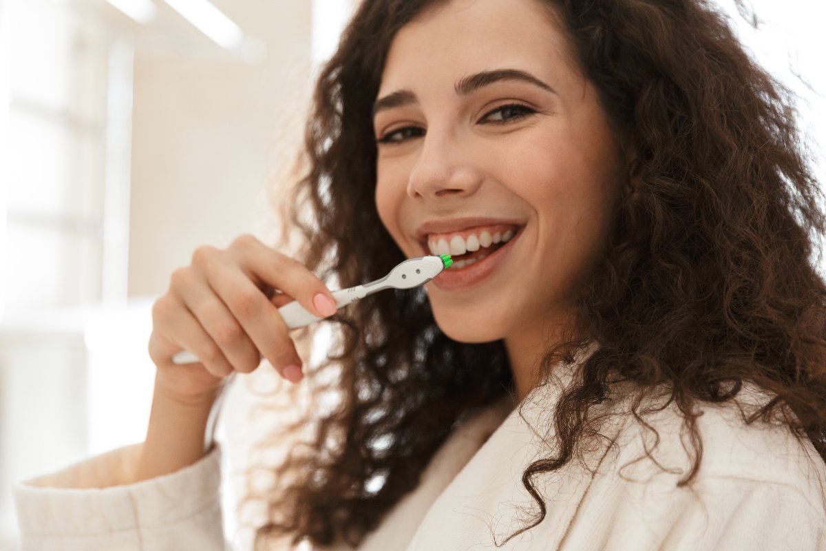 Woman brushing teeth in bright bathroom, smiling at camera during morning routine.