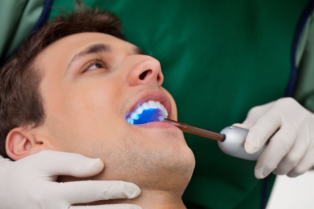 Patient receiving dental treatment with blue curing light while dentist works in clinical setting.