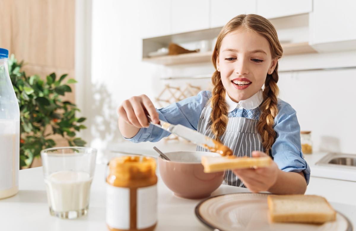 Young girl eating cereal at kitchen table with milk and bread nearby.