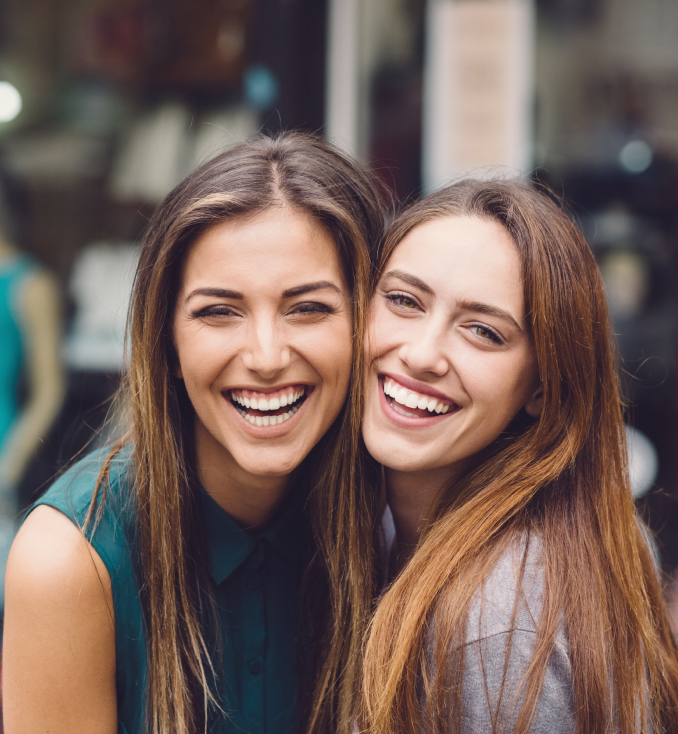 Two smiling women posing together outdoors in casual attire, urban background blurred.