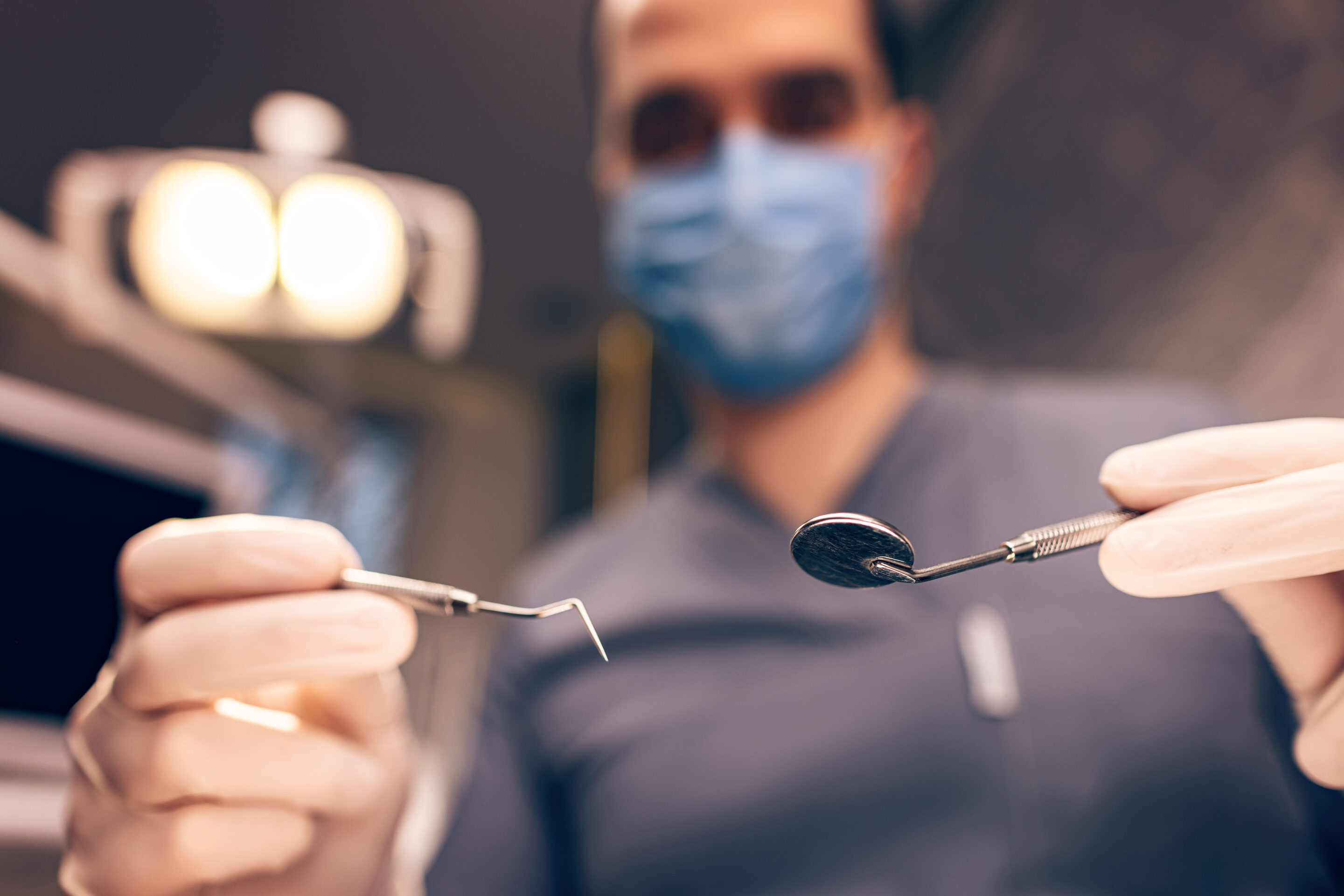 Dentist in mask holding dental tools in clinical setting with patient in background.